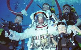 Scuba divers surrounding a man in an astronaut suit deep in a swimming pool.