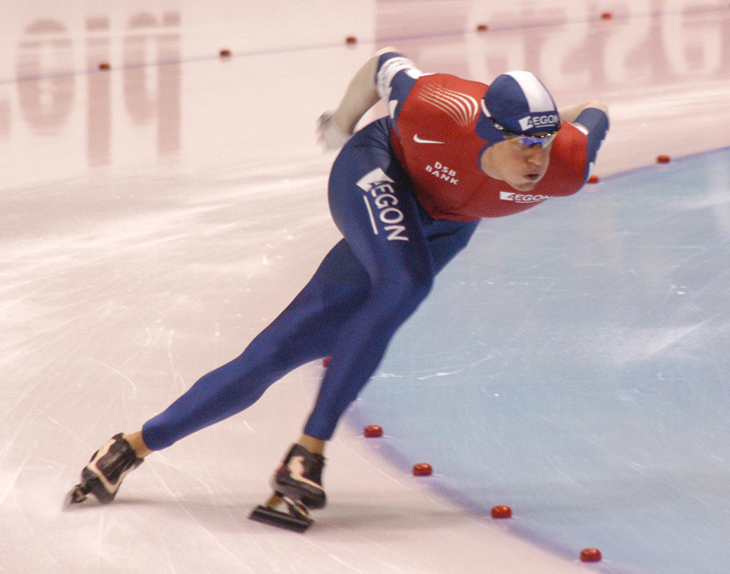A speed skater careens around a turn with head down, back arched, and arms back.