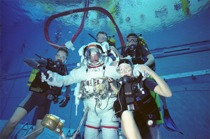Scuba divers surrounding a man in an astronaut suit deep in a swimming pool.